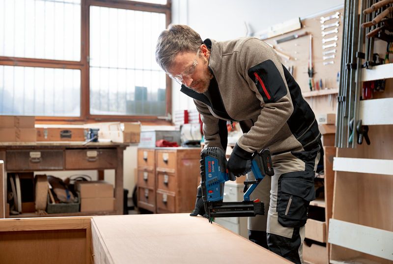 Eine Person, die Schutzausrüstung trägt, bedient einen akkubetriebenen Druckluftnagler an einem Holzschrank.
