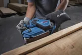 Person mit Sicherheitsausrüstung schleift Holz mit einem blauen Bandschleifer in einer Werkstatt.