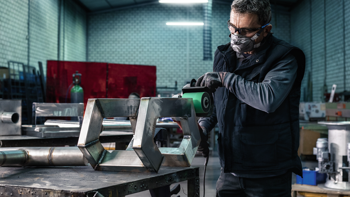 Eine Person mit Sicherheitsausrüstung schleift in einer Werkstatt eine Metallstruktur.
