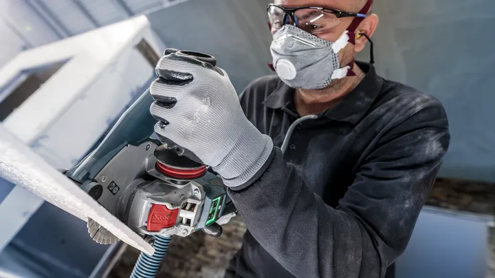 Eine Person mit Sicherheitsausrüstung schleift eine Oberfläche mit einem Elektrowerkzeug.