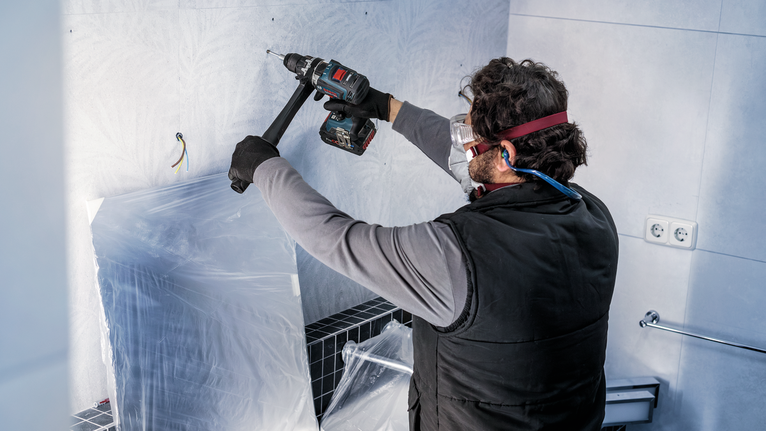 Eine Person mit Schutzausrüstung bohrt in eine geflieste Badezimmerwand.