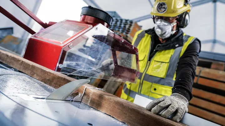 Eine Person mit Sicherheitsausrüstung führt Holz durch eine Tischkreissäge.