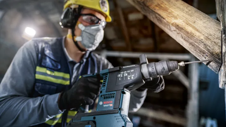 Eine Person mit Schutzausrüstung bohrt mit einem Elektrowerkzeug in einen Holzbalken.