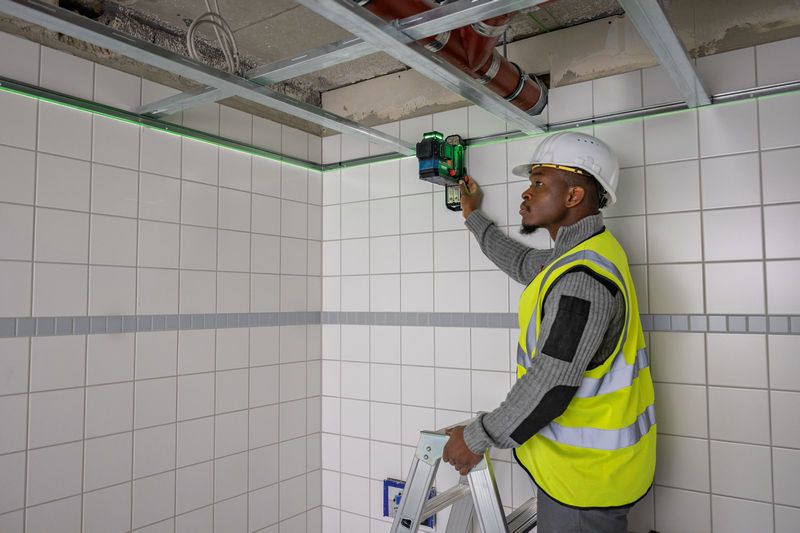 Eine Person mit Sicherheitsausrüstung richtet von einer Leiter aus ein Lasernivelliergerät auf einer gefliesten Wand aus.