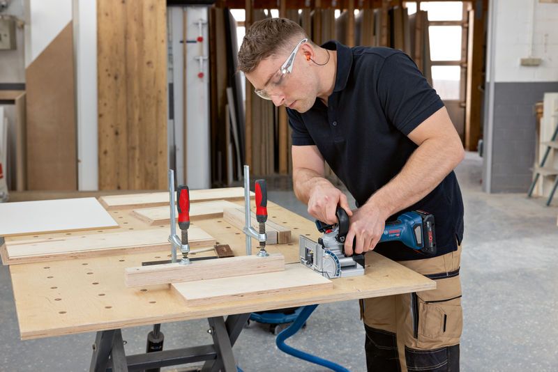 Eine Person mit Sicherheitsausrüstung verwendet eine Lamellofräse an eingespannten Holzstücken.