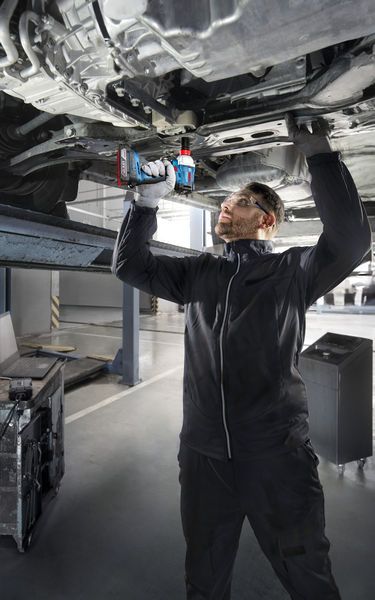 Eine Person mit Sicherheitsausrüstung verwendet eine Akku-Bohrmaschine unter einem angehobenen Fahrzeug.