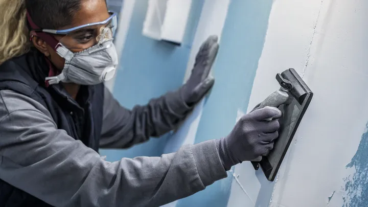 Person mit Sicherheitsausrüstung beim Schleifen einer Wand.