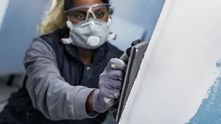 Eine Person mit Sicherheitsausrüstung schleift eine Wand mit einem Handschleifer.