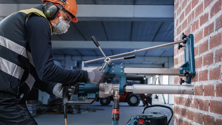 Eine Person mit Schutzausrüstung bohrt mit einem montierten Werkzeug in eine Ziegelwand.