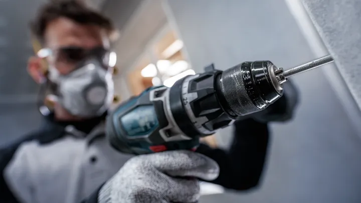 Person mit Sicherheitsausrüstung bohrt mit einem Elektrowerkzeug in eine Wand.