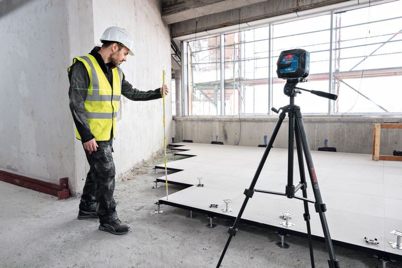 Eine Person mit Sicherheitsausrüstung nivelliert den Boden mithilfe eines Lasernivelliergeräts auf einem Stativ.
