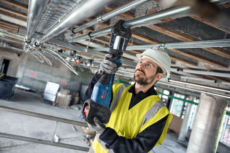 Eine Person mit Sicherheitsausrüstung verpresst eine Rohrverbindung mit einem Akku-Presswerkzeug über dem Kopf.