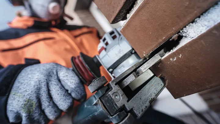 Eine Person mit Sicherheitsausrüstung schneidet mit einem Elektrowerkzeug eine braune Fliese.