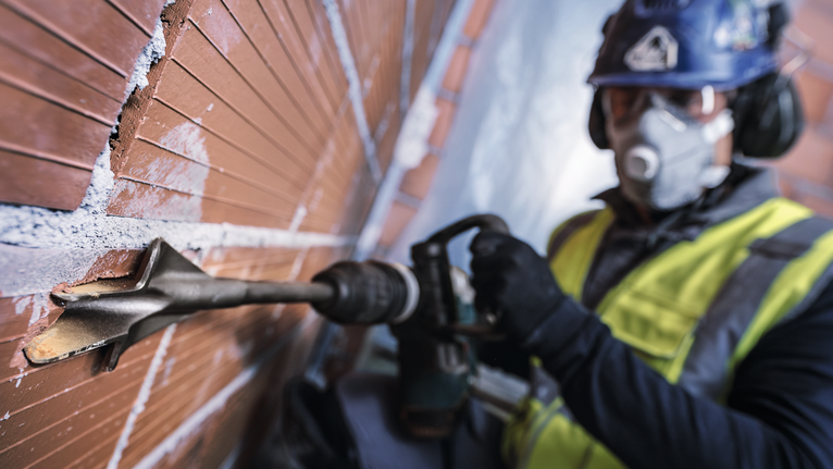 Person mit Schutzausrüstung bohrt mit Bohrhammer in eine Ziegelwand.