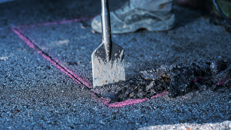 Eine Person mit Sicherheitsausrüstung bricht mit einem Meißelwerkzeug Asphalt entlang einer markierten Linie auf.
