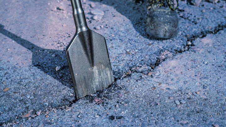Meißel bricht Asphalt, in der Nähe befindet sich eine Person mit Schutzausrüstung.