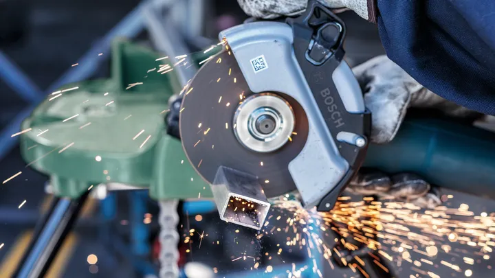 Eine Person mit Sicherheitsausrüstung schneidet mit einer Kreissäge durch ein Metallrohr, wobei Funken entstehen.