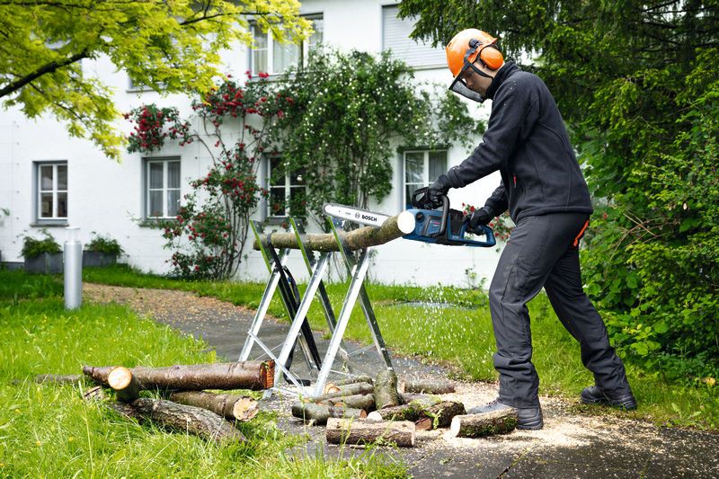 Eine Person mit Sicherheitsausrüstung verwendet eine Akku-Kettensäge, um im Freien Baumstämme zu schneiden.