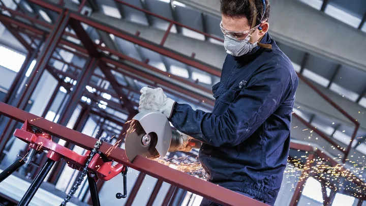 Eine Person mit Sicherheitsausrüstung schneidet mit einem Winkelschleifer ein Metallrohr, Funken fliegen.
