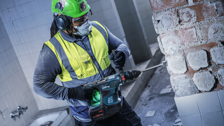 Eine Person mit Sicherheitsausrüstung bohrt in einer Baustelle in eine Ziegelwand.