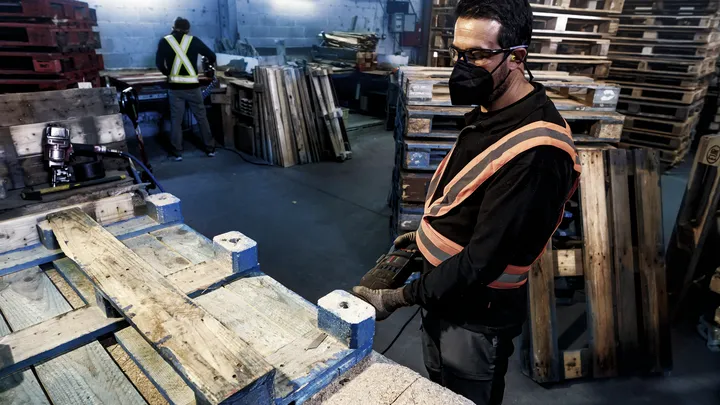 Eine Person mit Sicherheitsausrüstung repariert mit einem Elektrowerkzeug eine Holzpalette.