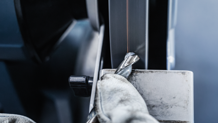 Person mit Sicherheitsausrüstung schärft einen Metallbohrer auf einer Doppelschleifmaschine.