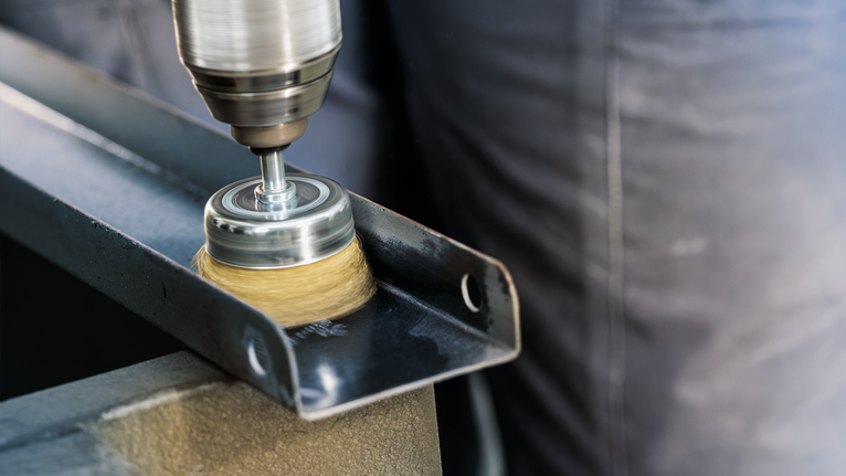 Eine Person poliert einen Metallträger mit einem Drahtbürstenaufsatz auf einer Bohrmaschine.