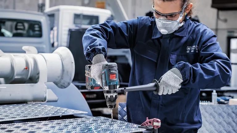 Eine Person mit Sicherheitsausrüstung bohrt in einer Industrieumgebung in eine Metalloberfläche.