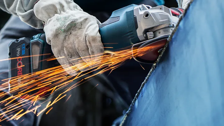 Eine Person mit Schutzausrüstung schleift Metall mit einem Elektrowerkzeug und erzeugt dabei Funken.