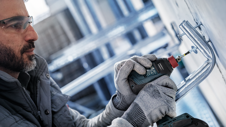 Person mit Schutzausrüstung bohrt in eine Metallwandhalterung.