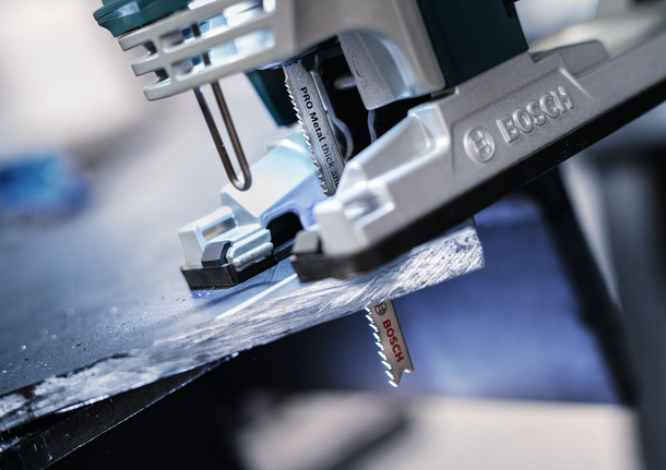 Eine Stichsäge schneidet in einer Werkstatt durch ein dunkles Metallblech.