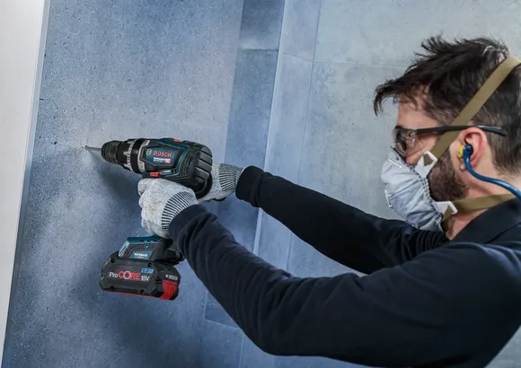 Person mit Schutzausrüstung bohrt in eine geflieste Wand.