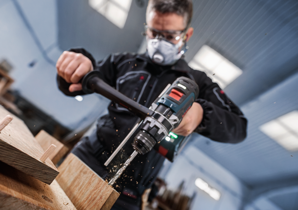 Person mit Sicherheitsausrüstung bohrt in Holz im Innenbereich.