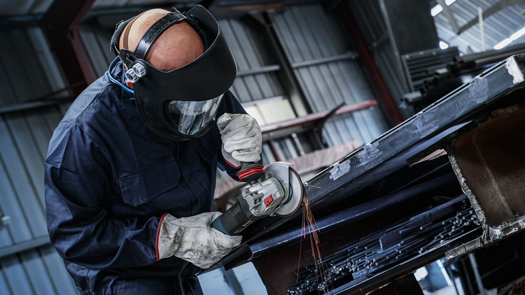 Eine Person mit Sicherheitsausrüstung schleift in einer industriellen Umgebung Metall und erzeugt dabei Funken.
