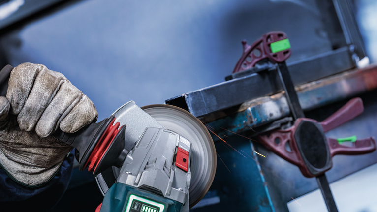 Eine Person mit Schutzausrüstung schleift einen Metallbalken und erzeugt dabei Funken.