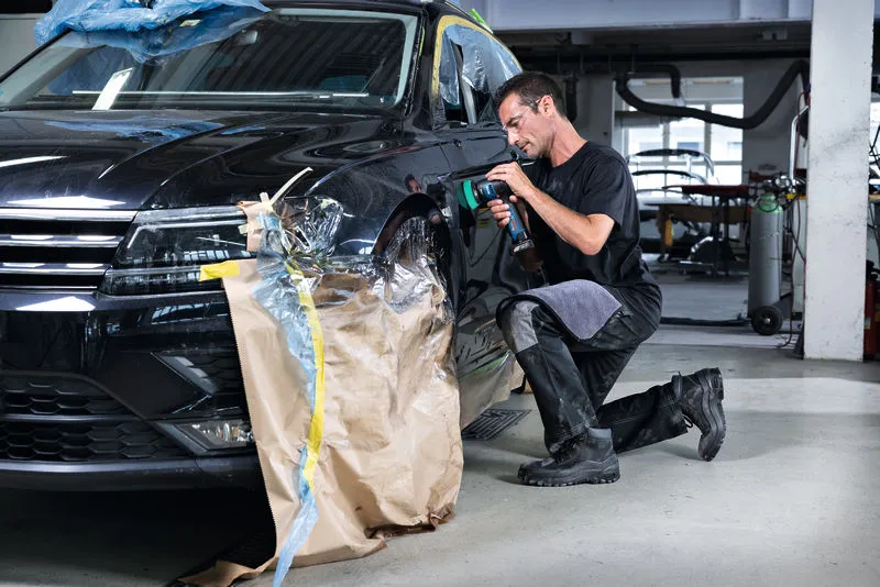 Eine Person mit Sicherheitsausrüstung verwendet in einer Autowerkstatt eine Poliermaschine an einem Kotflügel.