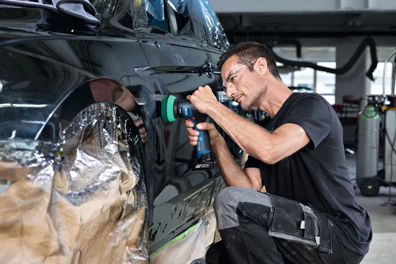 Eine Person mit Schutzausrüstung poliert eine Autotür mit einem Akkugerät.