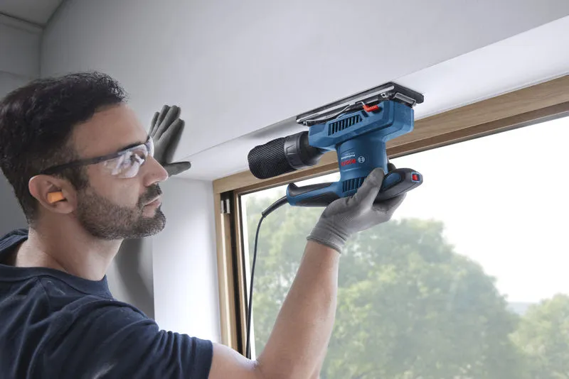 Eine Person mit Schutzausrüstung schleift einen Fensterrahmen mit einem Schwingschleifer.