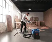 Eine Person mit Sicherheitsausrüstung verwendet in einer Holzwerkstatt einen Nass-Trocken-Sauger.