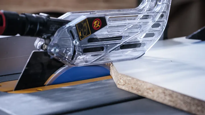 Eine Tischkreissäge schneidet mit montierter Schutzhaube durch ein Holzbrett.