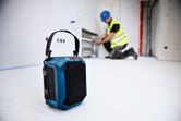 Ein tragbarer Baustellenlautsprecher liegt auf dem Boden, im Hintergrund arbeitet eine Person in Schutzausrüstung.
