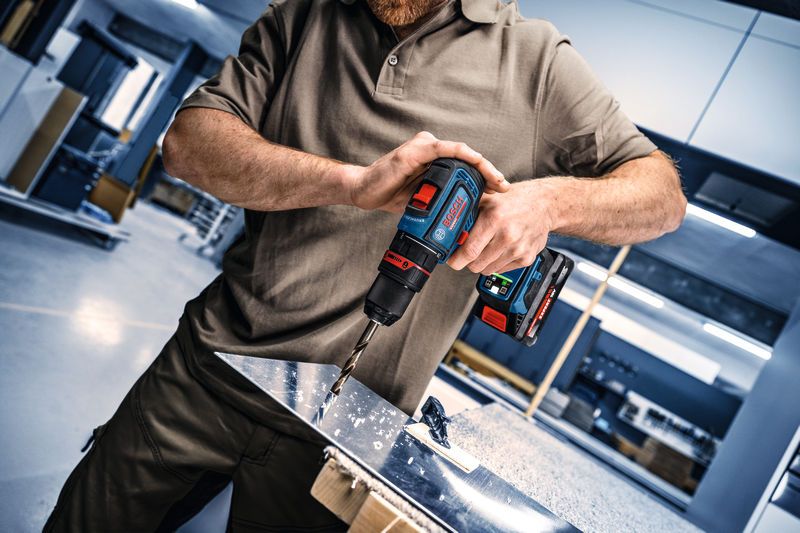 Person, die mit einem Akku-Bohrschrauber ein Loch in ein glänzendes Metallblech bohrt.