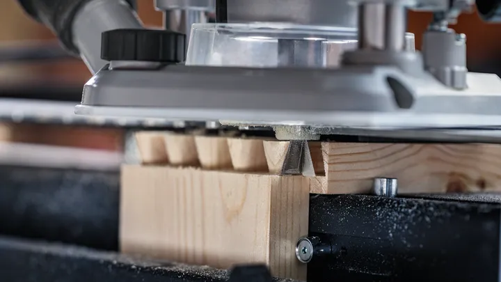 Mit einer Oberfräse werden Schwalbenschwanzverbindungen in ein Stück helles Holz geschnitzt.