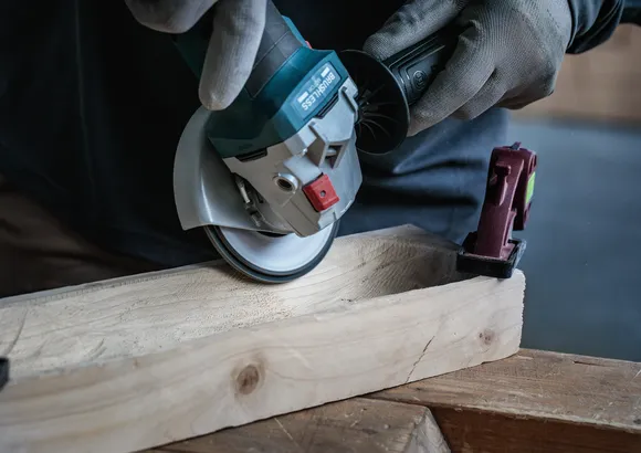 Eine Person mit Schutzausrüstung schleift ein Holzbrett mit einem Winkelschleifer.