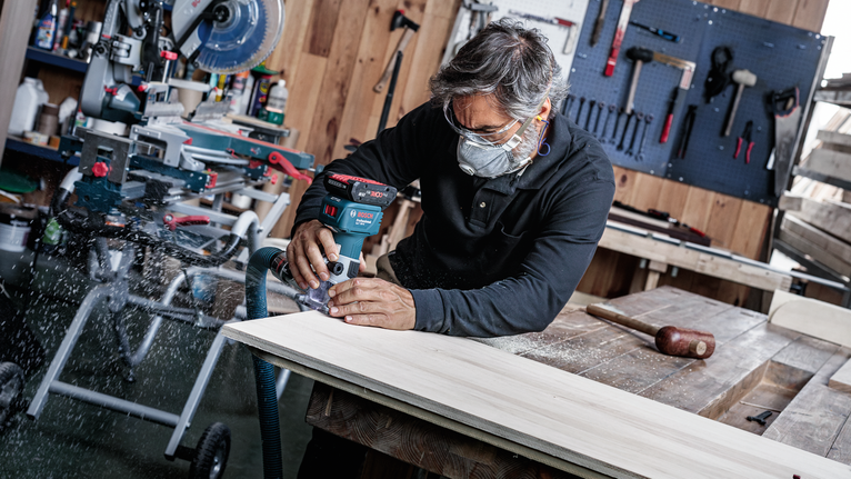Person mit Schutzausrüstung, die einen Akku-Fräser auf einem Holzbrett verwendet.