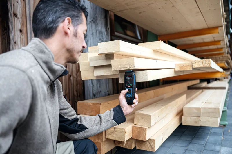Eine Person misst die Feuchtigkeit in gestapeltem Holz mit einem digitalen Materialfeuchtemessgerät.