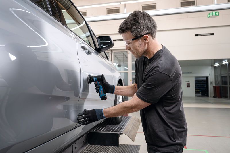 Eine Person mit Sicherheitsausrüstung schleift eine Autotür mit einem Exzenterschleifer.