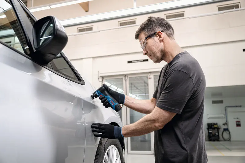 Eine Person mit Schutzausrüstung schleift mit einem Schwingschleifer eine Autotür.