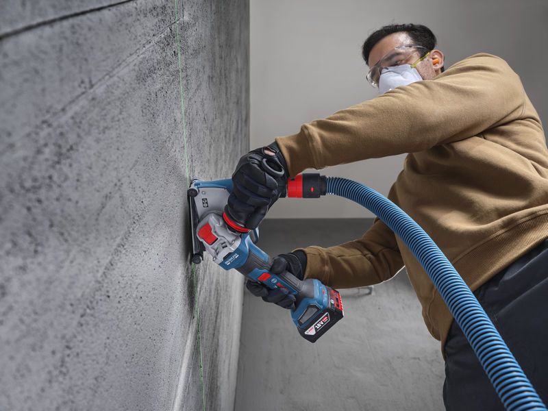 Eine Person mit Schutzausrüstung schleift mit einem Akku-Winkelschleifer eine Betonwand.