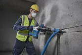 Eine Person, die Schutzausrüstung trägt, bohrt mit einem Elektrowerkzeug in eine Betonwand.